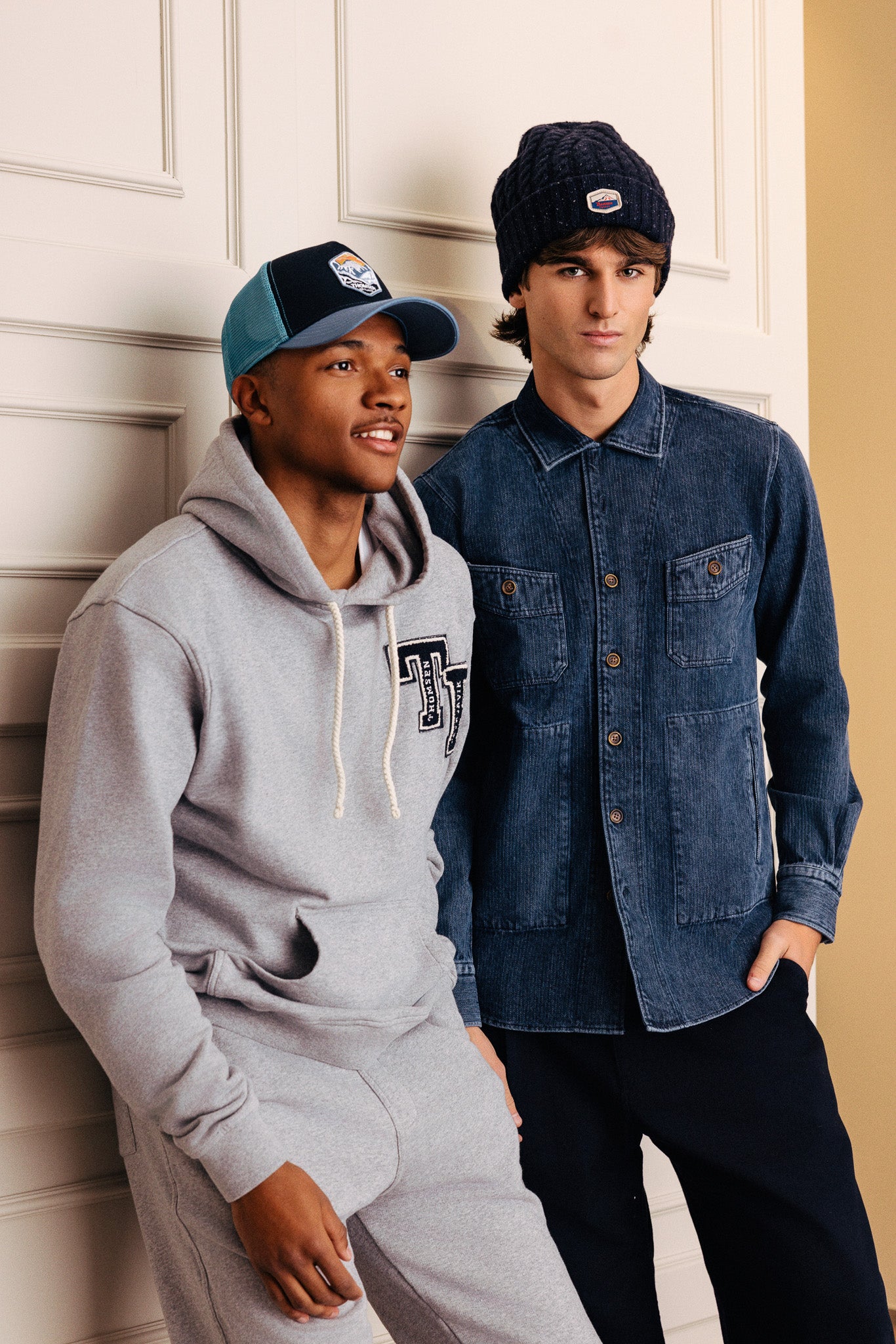 Tveir ungir menn. Annar í hettupeysu og íþróttabuxum með derhúfu. Hinn er í yfirskyrtu og með ullarhúfu.