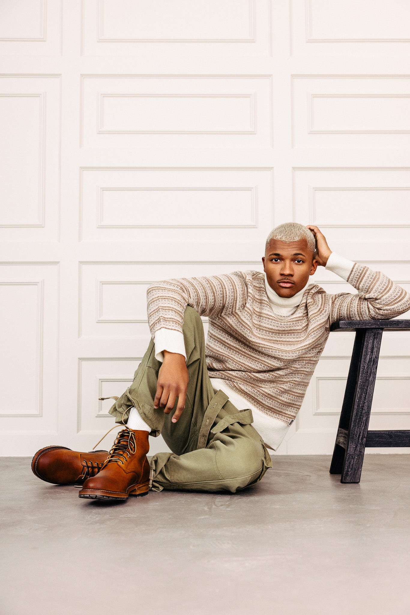 Módelið situr á gólfinu. Er í rúllukragapeysu úr ull, grænum buxum og brúnum leðurskóm.