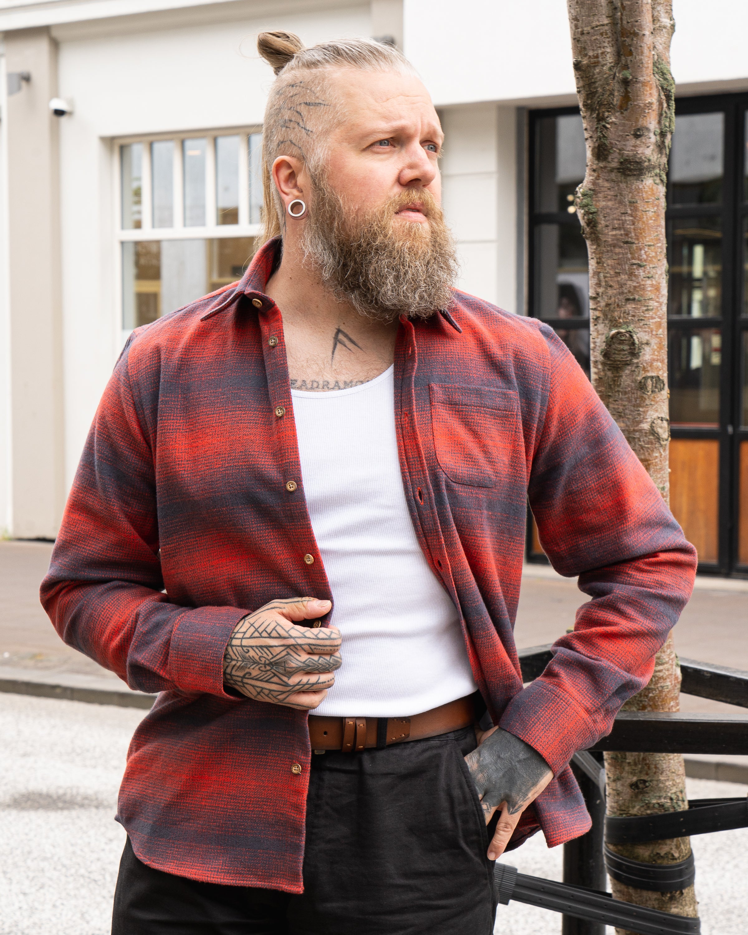 Man wearing a grey and red check Kári shirt over a white tank top, showcasing a casual style with a beard and tattoos.